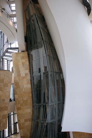 A modern architectural interior with large, curved glass panels and smooth, flowing walls. The design showcases a blend of metal, glass, and stone elements with natural light filtering through expansive windows. The space appears open and airy, with a focus on geometric and organic forms.