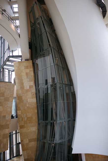 A modern architectural interior with large, curved glass panels and smooth, flowing walls. The design showcases a blend of metal, glass, and stone elements with natural light filtering through expansive windows. The space appears open and airy, with a focus on geometric and organic forms.