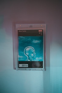 A framed comic book cover depicting a futuristic, stylized character with an oversized head in profile view. The background is blue with a cloud-like texture. The comic is a Marvel publication, specifically the first issue of Silver Surfer.