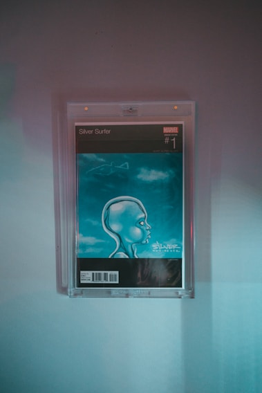 A framed comic book cover depicting a futuristic, stylized character with an oversized head in profile view. The background is blue with a cloud-like texture. The comic is a Marvel publication, specifically the first issue of Silver Surfer.