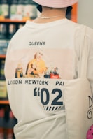 A person is seen from behind wearing a graphic t-shirt with an image featuring a person in a bright yellow shirt and various drink bottles, including Coca-Cola. The shirt has the word 'QUEENS' and locations like London, New York, and Paris printed on it, along with the number '02'. The person also carries a tote bag and wears a light pink hat.