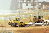 A construction site with heavy machinery, including a yellow steamroller and a bulldozer, surrounded by large concrete pipes. There is fencing around the area with various construction materials scattered. Buildings and cranes are visible in the background, suggesting ongoing construction activities.