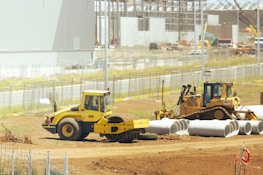 A construction site with heavy machinery, including a yellow steamroller and a bulldozer, surrounded by large concrete pipes. There is fencing around the area with various construction materials scattered. Buildings and cranes are visible in the background, suggesting ongoing construction activities.