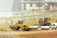 A construction site with heavy machinery, including a yellow steamroller and a bulldozer, surrounded by large concrete pipes. There is fencing around the area with various construction materials scattered. Buildings and cranes are visible in the background, suggesting ongoing construction activities.