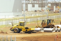 A construction site with heavy machinery, including a yellow steamroller and a bulldozer, surrounded by large concrete pipes. There is fencing around the area with various construction materials scattered. Buildings and cranes are visible in the background, suggesting ongoing construction activities.