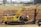 A large yellow excavator with the brand name 'Azzona' on the arm is positioned on a construction site with dirt and various construction materials. In the background, there is a mix of industrial buildings and vehicles, including white cars and trucks.