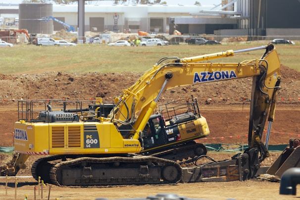 A large yellow excavator with the brand name 'Azzona' on the arm is positioned on a construction site with dirt and various construction materials. In the background, there is a mix of industrial buildings and vehicles, including white cars and trucks.