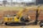 A large yellow excavator with the brand name 'Azzona' on the arm is positioned on a construction site with dirt and various construction materials. In the background, there is a mix of industrial buildings and vehicles, including white cars and trucks.