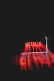 A glowing laptop screen displaying the word 'Netflix' against a dark background. The red text appears prominently, casting a reflection or light effect across the image.