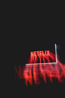 A glowing laptop screen displaying the word 'Netflix' against a dark background. The red text appears prominently, casting a reflection or light effect across the image.