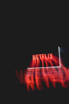 A glowing laptop screen displaying the word 'Netflix' against a dark background. The red text appears prominently, casting a reflection or light effect across the image.