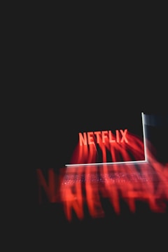 A glowing laptop screen displaying the word 'Netflix' against a dark background. The red text appears prominently, casting a reflection or light effect across the image.