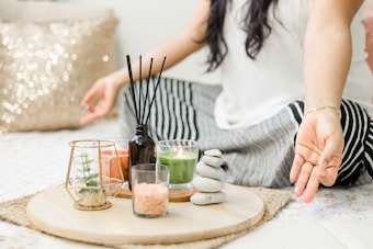 A person seated cross-legged on a bed or mat, engaging in meditation or relaxation. To their side, a wooden tray holds various decorative items, including a small plant in a glass holder, candles, a black diffuser with reeds, a small bowl of pink salt, and a stack of smooth stones. The setting conveys a calm and peaceful ambiance, enhanced by soft lighting and a glittery pillow in the background.