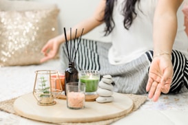 A person seated cross-legged on a bed or mat, engaging in meditation or relaxation. To their side, a wooden tray holds various decorative items, including a small plant in a glass holder, candles, a black diffuser with reeds, a small bowl of pink salt, and a stack of smooth stones. The setting conveys a calm and peaceful ambiance, enhanced by soft lighting and a glittery pillow in the background.