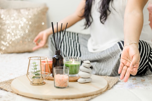 A person seated cross-legged on a bed or mat, engaging in meditation or relaxation. To their side, a wooden tray holds various decorative items, including a small plant in a glass holder, candles, a black diffuser with reeds, a small bowl of pink salt, and a stack of smooth stones. The setting conveys a calm and peaceful ambiance, enhanced by soft lighting and a glittery pillow in the background.