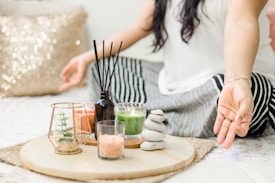 A person seated cross-legged on a bed or mat, engaging in meditation or relaxation. To their side, a wooden tray holds various decorative items, including a small plant in a glass holder, candles, a black diffuser with reeds, a small bowl of pink salt, and a stack of smooth stones. The setting conveys a calm and peaceful ambiance, enhanced by soft lighting and a glittery pillow in the background.