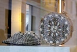 A pair of intricately patterned slippers and a spherical object are displayed together. The slippers feature a black and white geometric design embellished with cowrie shells along the edges. The spherical object is decorated with a detailed arrangement of shells and beads in concentric patterns.