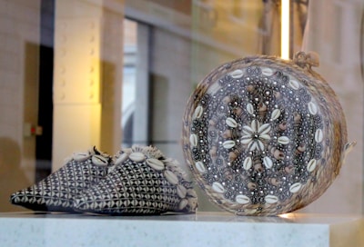 A pair of intricately patterned slippers and a spherical object are displayed together. The slippers feature a black and white geometric design embellished with cowrie shells along the edges. The spherical object is decorated with a detailed arrangement of shells and beads in concentric patterns.