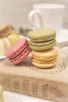 A stack of pastel macarons in soft pink, beige, and cream tones arranged on a light wooden table with a cup of coffee.