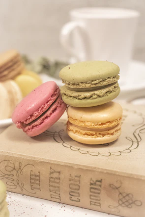 A stack of pastel macarons in soft pink, beige, and cream tones arranged on a light wooden table with a cup of coffee.