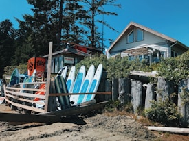 A coastal scene with a rustic shop surrounded by surfboards placed against a wooden fence. The shop, adorned with signs including a 'CLOSED' sign, sits next to a house with a high roof. Tall trees and greenery frame the setting, emphasizing a natural, serene environment.