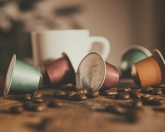 Several colorful coffee capsules are scattered on a wooden surface alongside coffee beans. In the blurred background, a white ceramic coffee mug is visible, creating a cozy and inviting atmosphere.
