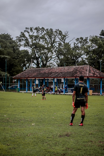 A group of soccer players are on a grassy field with a team building and trees in the background. One player in the foreground is wearing gloves and a jersey with the number 1, standing ready on the field. Other players are visible near the building, either standing or sitting.