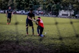 Two soccer players are on a grassy field; one is helping another off the ground. They are wearing different colored jerseys, indicating they might be from opposing teams or in training. Other players and some parked cars are visible in the background, suggesting an urban setting.