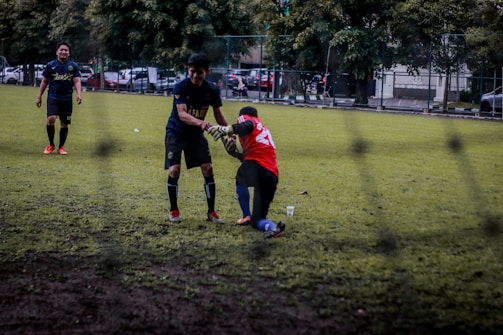 Two soccer players are on a grassy field; one is helping another off the ground. They are wearing different colored jerseys, indicating they might be from opposing teams or in training. Other players and some parked cars are visible in the background, suggesting an urban setting.