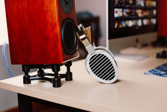 On a wooden desk, there is a red wooden speaker on a black stand and a pair of headphones resting against it. In the background, a computer monitor displays a colorful image with multiple small windows.