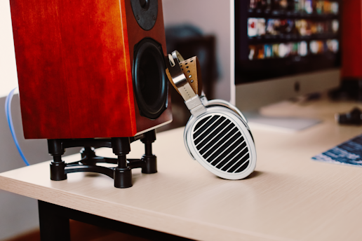 On a wooden desk, there is a red wooden speaker on a black stand and a pair of headphones resting against it. In the background, a computer monitor displays a colorful image with multiple small windows.