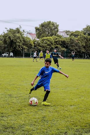 A young boy in a blue soccer jersey and black shorts is kicking a soccer ball on a grassy field. In the background, several other players, some wearing yellow bibs, are actively engaged in the game, with lush green trees and some buildings visible beyond the field.