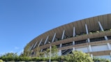 A modern architectural structure with a distinctive wooden slatted design forms the upper part of a large building. Green plants are integrated into the design, visible on the ledges and around the structure. A few trees and shrubbery are present in front of the building, alongside a clear blue sky.
