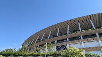 A modern architectural structure with a distinctive wooden slatted design forms the upper part of a large building. Green plants are integrated into the design, visible on the ledges and around the structure. A few trees and shrubbery are present in front of the building, alongside a clear blue sky.