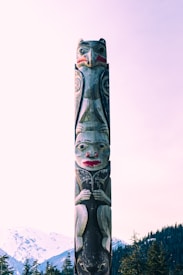 A traditional totem pole stands prominently outdoors, intricately carved with symbolic figures and patterns. The pole features vibrant colors, with the main figures possibly representing animals or spirits. Snow-covered mountains and evergreen trees fill the background, adding a natural and serene setting.