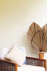 A cozy seating area with a woven rattan chair featuring a beige cushion and decorative pillows. A large dried palm leaf in a vase provides an earthy accent against a neutral wall.