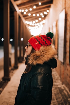 A person wearing a red knit hat with a pom-pom and a thick winter coat with a fur-lined hood stands under a row of glowing string lights. The setting appears to be outdoors with a brick wall in the background, and there is a cozy ambiance created by the warm lighting.