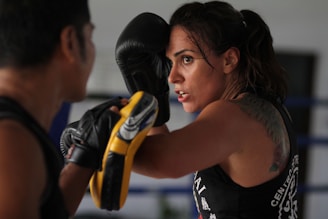 A woman in intense focus engages in boxing training, with gloves and protective gear. Her tattoo is partially visible on her shoulder, and she appears determined as she spars with a partner.