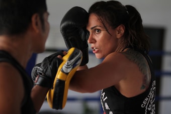 A woman in intense focus engages in boxing training, with gloves and protective gear. Her tattoo is partially visible on her shoulder, and she appears determined as she spars with a partner.