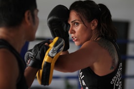A woman in intense focus engages in boxing training, with gloves and protective gear. Her tattoo is partially visible on her shoulder, and she appears determined as she spars with a partner.