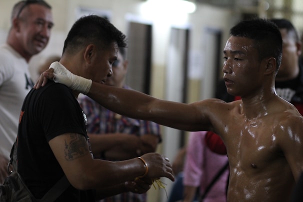 A male boxer, visibly sweaty, stands facing another person with his arm resting on the person's shoulder. The dimly lit setting suggests a gym or training area, with other observers in the background.