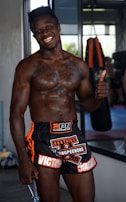 A muscular man smiling confidently, wearing black and orange sports shorts with visible tattoos on his chest. He is giving a thumbs-up gesture while standing in a gym environment with a punching bag in the background.