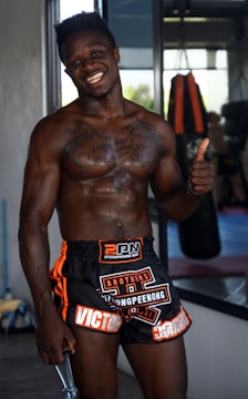 A muscular man smiling confidently, wearing black and orange sports shorts with visible tattoos on his chest. He is giving a thumbs-up gesture while standing in a gym environment with a punching bag in the background.