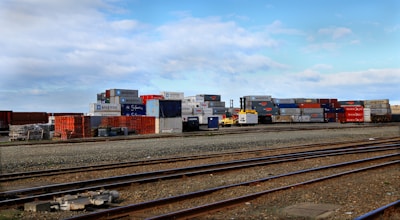 Stacks of colorful shipping containers are piled next to a set of railway tracks. The containers bear branding from various shipping companies, and the area appears to be part of a shipping yard or freight terminal. The sky is clear with some scattered clouds.