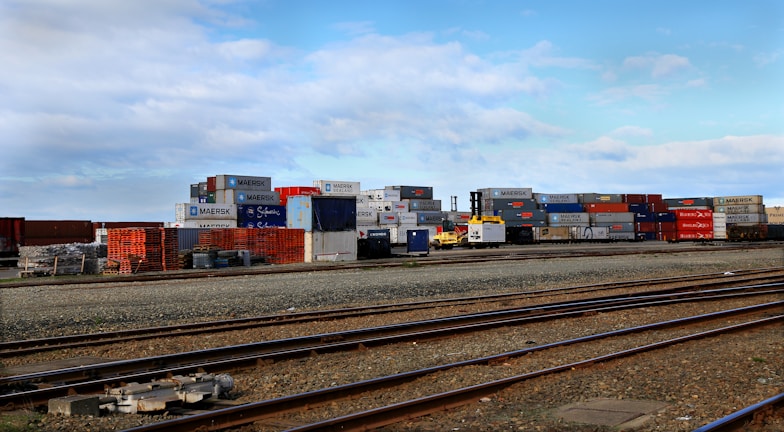 Stacks of colorful shipping containers are piled next to a set of railway tracks. The containers bear branding from various shipping companies, and the area appears to be part of a shipping yard or freight terminal. The sky is clear with some scattered clouds.