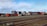 Stacks of colorful shipping containers are piled next to a set of railway tracks. The containers bear branding from various shipping companies, and the area appears to be part of a shipping yard or freight terminal. The sky is clear with some scattered clouds.