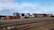 Stacks of colorful shipping containers are piled next to a set of railway tracks. The containers bear branding from various shipping companies, and the area appears to be part of a shipping yard or freight terminal. The sky is clear with some scattered clouds.
