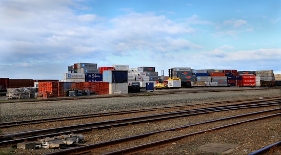 Stacks of colorful shipping containers are piled next to a set of railway tracks. The containers bear branding from various shipping companies, and the area appears to be part of a shipping yard or freight terminal. The sky is clear with some scattered clouds.