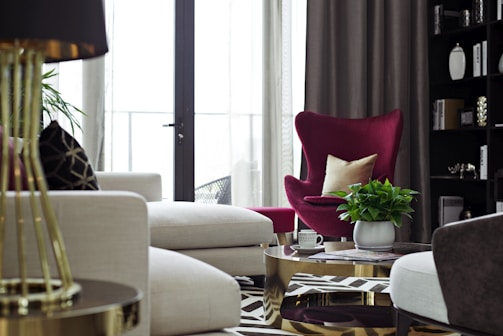 A modern living room with a chic interior design featuring a dark magenta armchair alongside cream-colored sofas. A small coffee table sits in front, topped with a green potted plant and a few small decorative items like cups. Curtains partly cover tall windows, allowing soft natural light into the space. To the right, a black shelving unit holds various ornaments and books, contributing to a sophisticated ambiance.