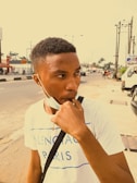 A young person with short hair is wearing a casual white T-shirt with blue lettering. They have white earphones in their ears and are holding a face mask near their chin, looking to the side. The background features an urban street setting with some cars, palm trees, and buildings visible.
